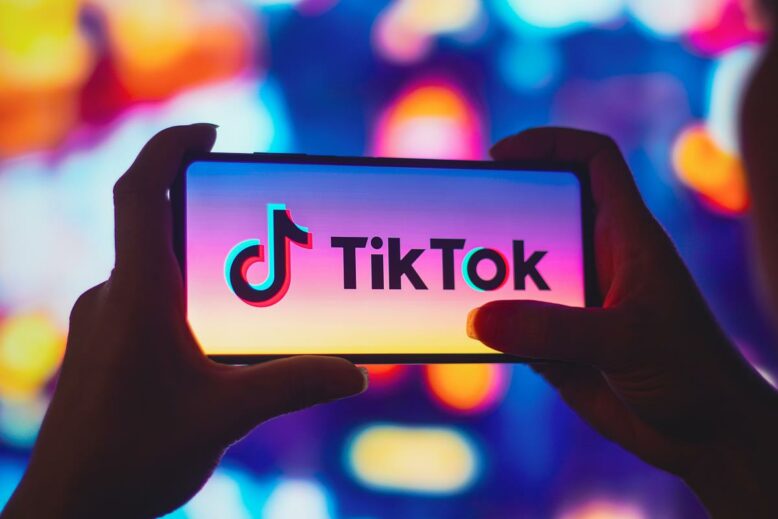 Smartphone mit TikTok-Logo auf dem Bildschirm, gehalten von zwei Händen vor einem unscharfen Hintergrund mit bunten Lichtern
